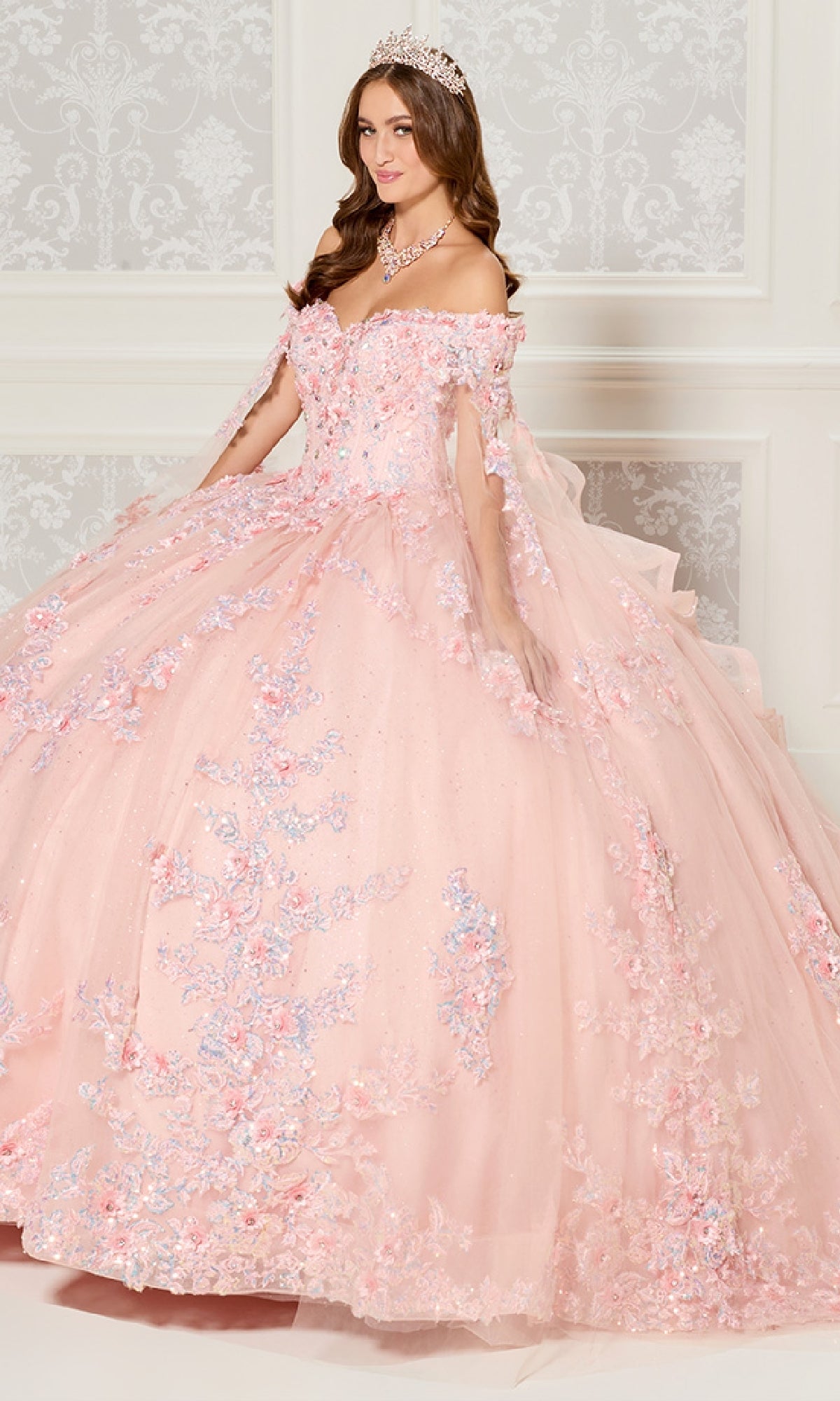 A woman in the Quinceanera Dress PR30116 By Princesa, featuring a pink off-the-shoulder design with floral details and a fitted corset bodice, wears a tiara as she stands before an ornate white wall.